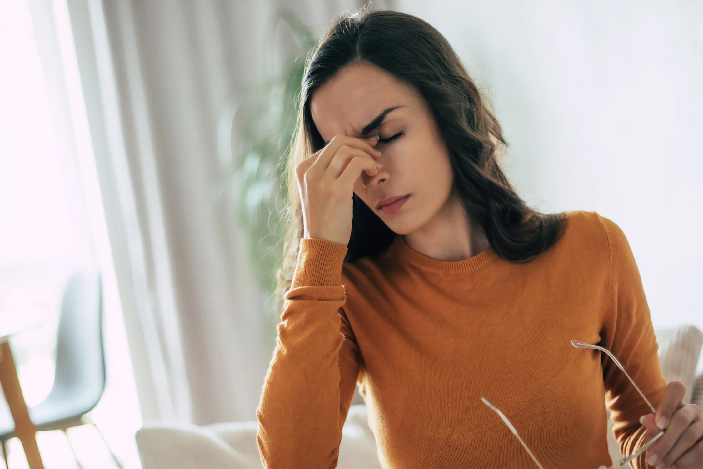A woman in an orange sweater sitting on a couch, holding her glasses and rubbing the bridge of her nose in visible stress or fatigue, symbolizing how stress can affect menstrual health.